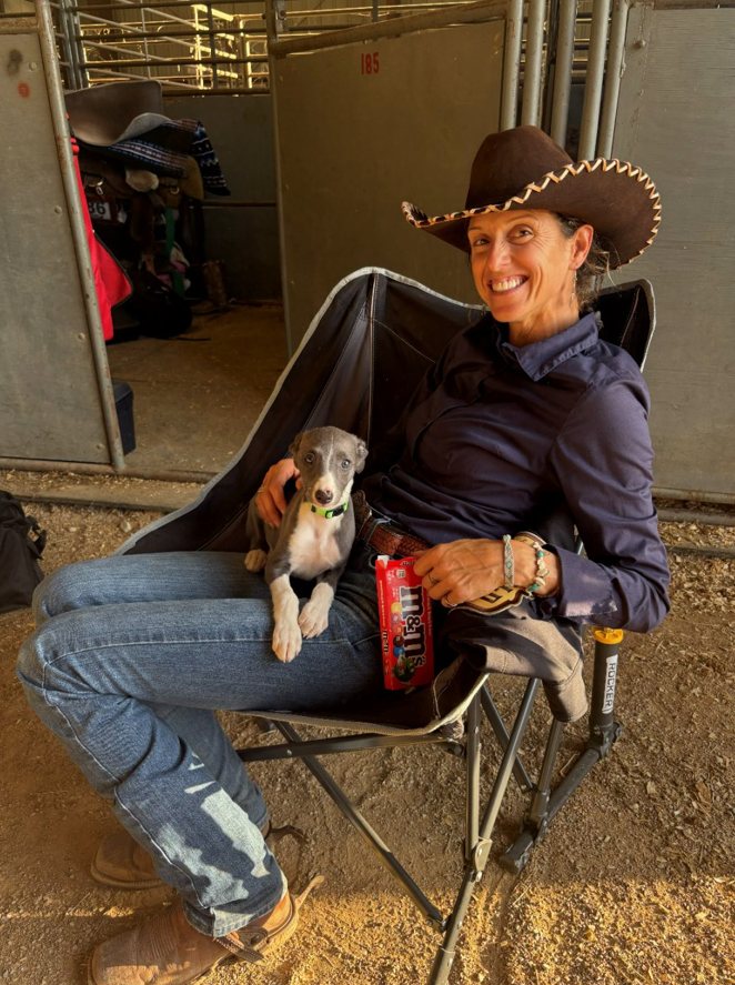 Trainer Tania Morton smiling in a cowboy hat, seated in a camp chair in the barn aisle with a whippet puppy on her lap